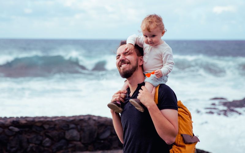 A man with a yellow rucksack carries a child by the sea in Melbourne’s west, sharing a playful moment—perfect for massage therapy.