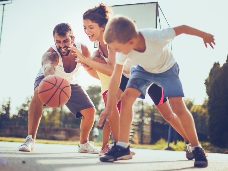 Two adults and a child play basketball outside in sunny Altona, Melbourne’s west—a great way to stay active before a sports massage.