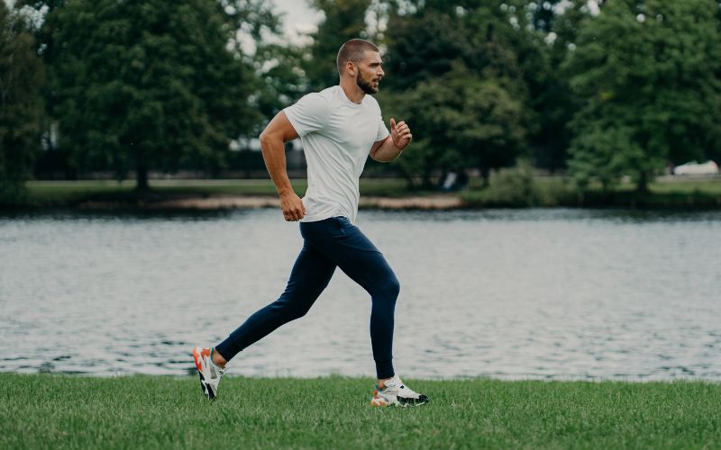 A man in sportswear jogs on the grass by the water in Melbourne’s west, trees behind him, enjoying a post-exercise run.
